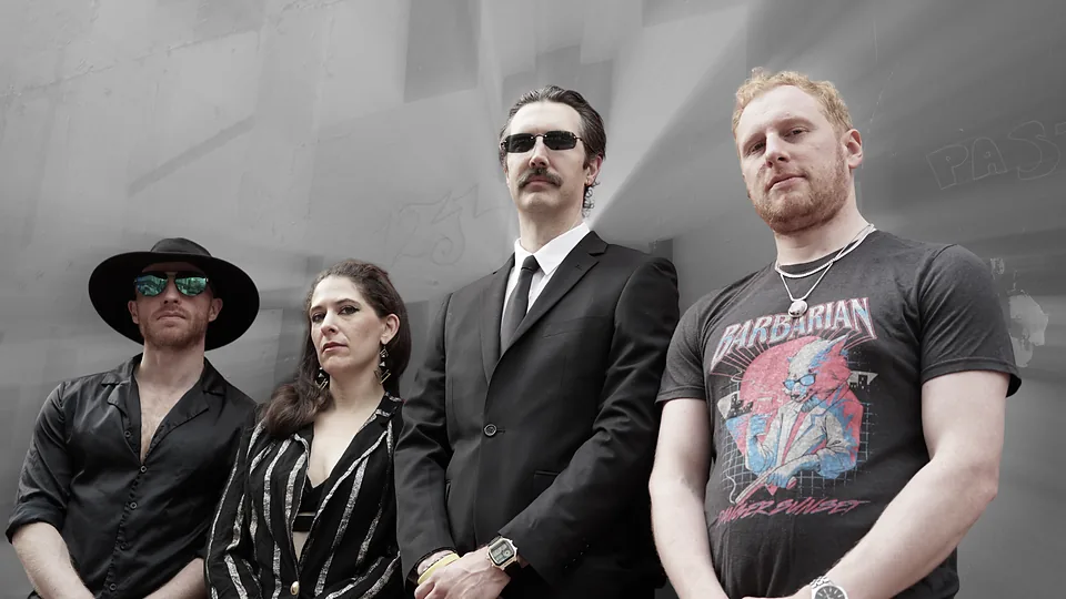 The four members of Ork standing shoulder to shoulder, in sharp focus against a grey, motion-blurred wall: one in a wide-brim hat and mirrored sunglasses, one in a striped jacket, one in a black suit and tie, one in a graphic T-shirt.